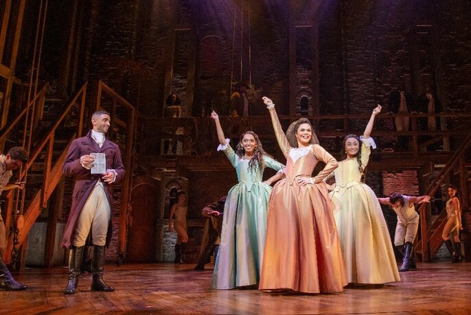 Three female cast members of Hamilton wearing crinolines, standing next to a man holding a declaration.