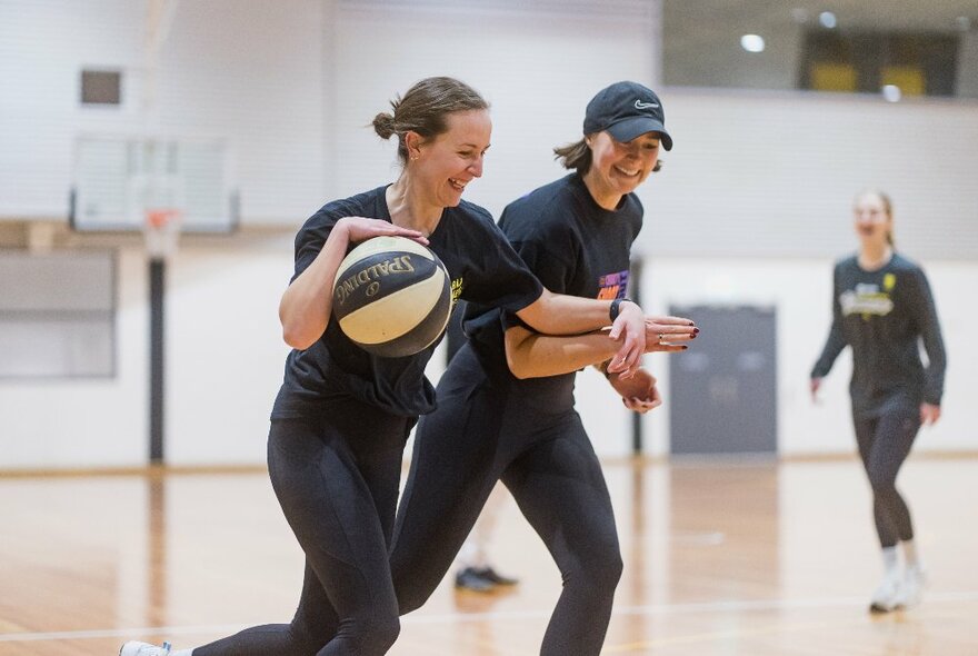 Two smiling women playing basketball inside a basketball stadium, with another woman in the background.