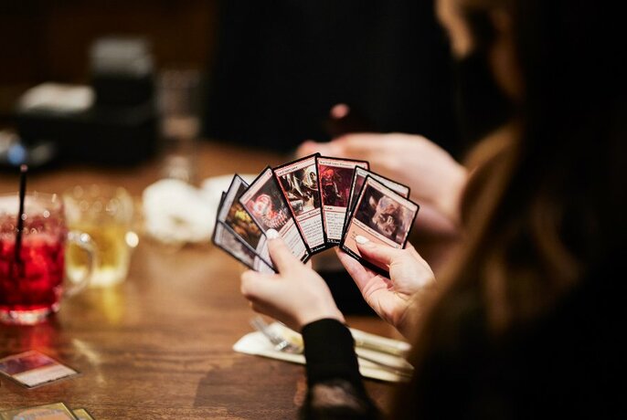 Hand of cards in hands, seen over player's shoulder; drinks on table to left.