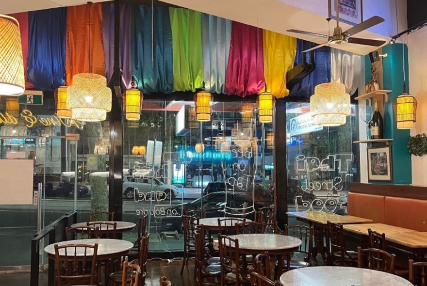 Interior of restaurant with round tables, chairs, longer booth seats and tables against a wall, brightly coloured wall hangings and a view through the window onto the street.
