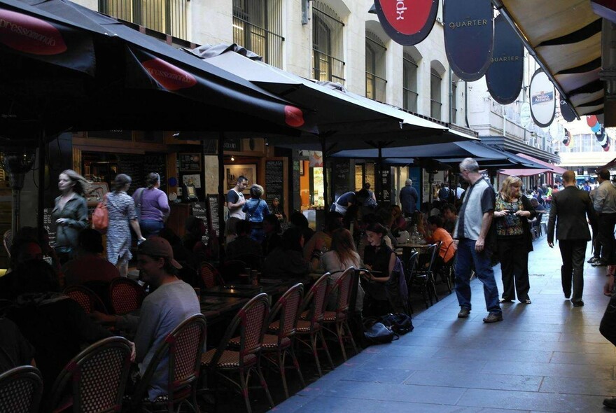 People walking past a Degraves Street outdoor cafe with lines of chairs under umbrellas.