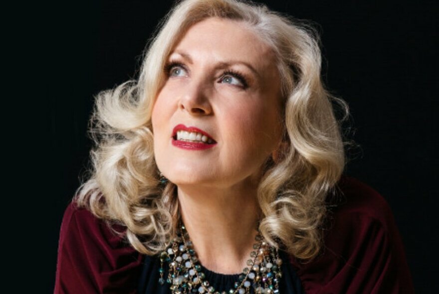 Singer Janet Ross-Fahy with curly blonde hair looking upwards against a black background. 