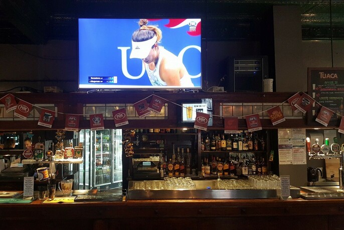 Hotel bar with rows of bottles and beer fridge, tennis screening on large screen in background.