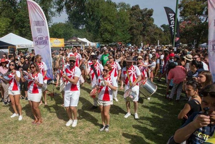 Rows of people in red and white outfits dancing and playing musical instruments in an outdoor setting with crowds watching them. 