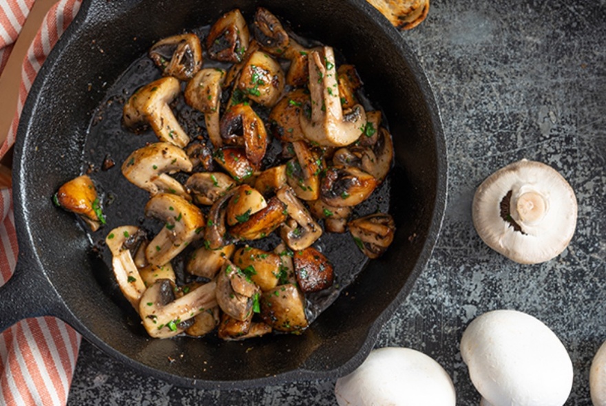 Looking straight down at a cast iron frying pan with browned mushrooms and herbs with scattered raw mushrooms beside it. 