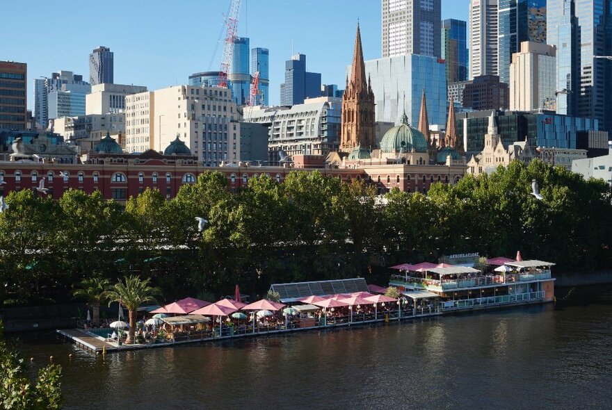 Arbory Afloat, a floating bar and restaurant on the Yarra RIver, with trees and the Melbourne city skyline visible behind it and the river in the foreground. 