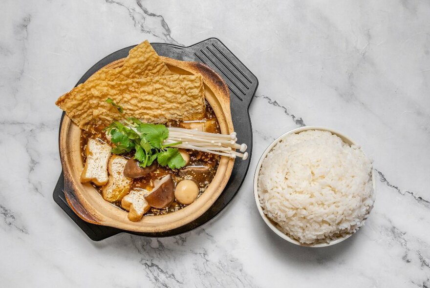 Overhead view of a claypot with a side dish of rice.