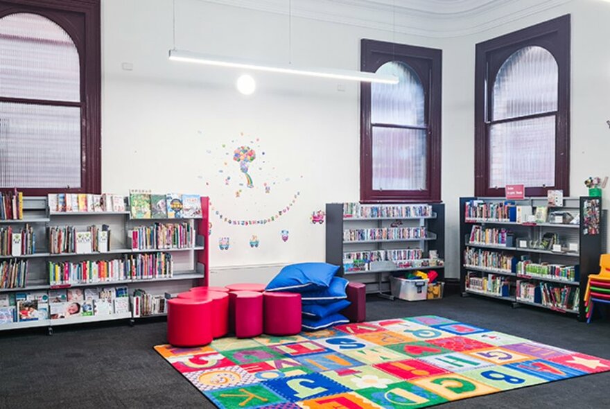 Brightly coloured rug, floor cushions and shelves of books in the interior of North Melbourne Library.