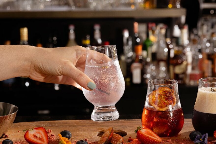 Hand picking up a cocktail in a glass from a bar counter, with bottles of alcohol visible in the background.