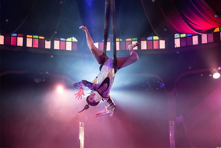 An acrobatic dancer hangs upside down by their legs during a performance with pink and purple lighting. 