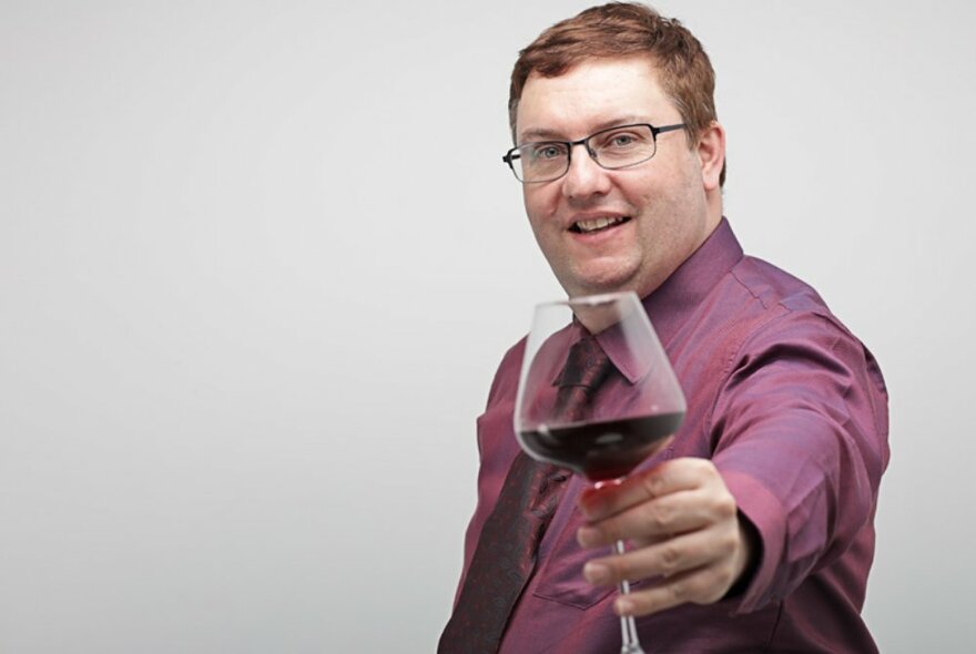 Middle-aged man wearing a purple shirt, holding out a glass of red wine in his hand, posed against a pale wall.