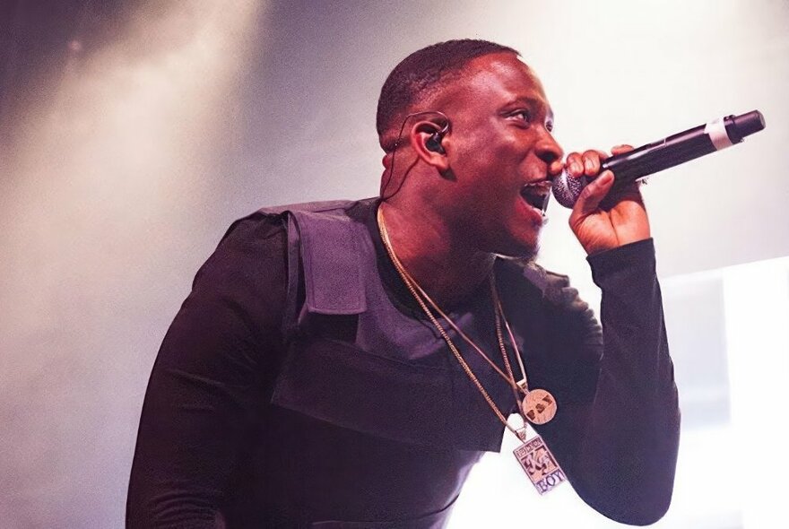 English musician, Kojo Funds, rapping into a microphone on stage, wearing all black and two statement necklaces.