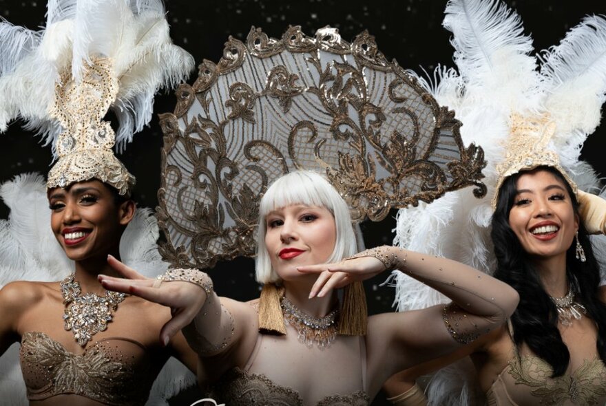 Three burlesque dancers wearing jewels and elaborate headdresses, gesticulating with their hands.