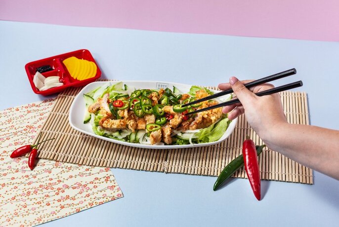 Orange table with a bowl of Korean noodles, bamboo mat, hands holding chopsticks and condiments.