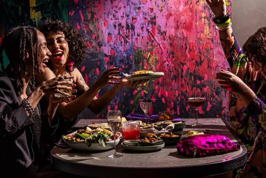 Three women seated at a table with plates of delicious food, laughing, in front of a brightly coloured abstract artwork on the wall.