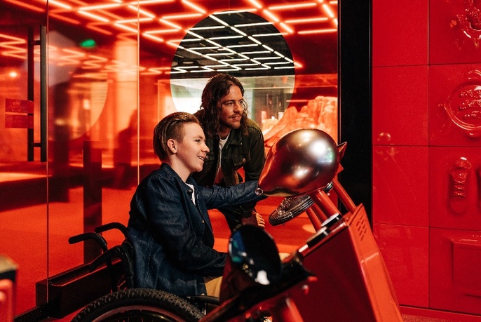 Two people (one in wheelchair, one crouched behind) in bright red room behind a console and looking ahead.