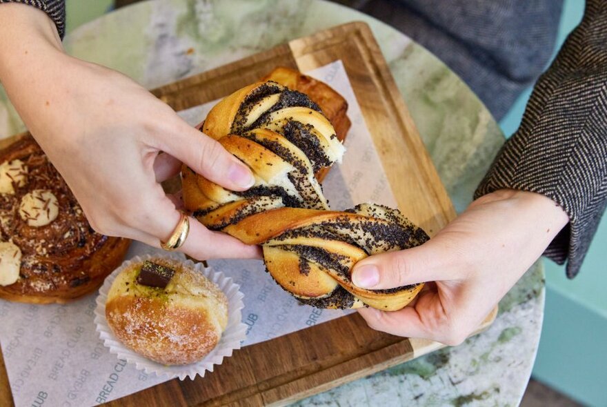 Pair of hands pulling apart a plaited pastry that is covered in poppy-seeds, above a wooden tray that contains other sweet pastries.