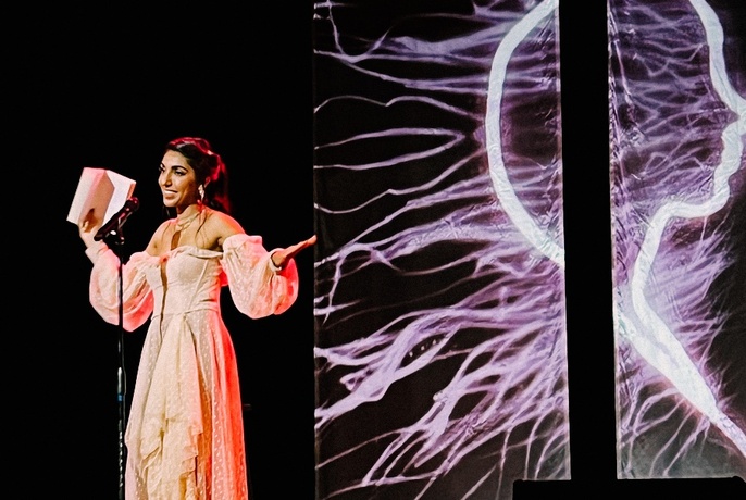 Poet Rupi Kaur smiling and wearing a long dress, holding a book in her hands, standing in front of a microphone on a stage.