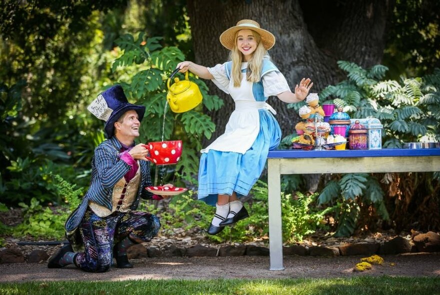 Performers dressed as The Mad Hatter and Alice in Wonderland acting out a scene where Alice pours tea from a teapot into an oversize cup.