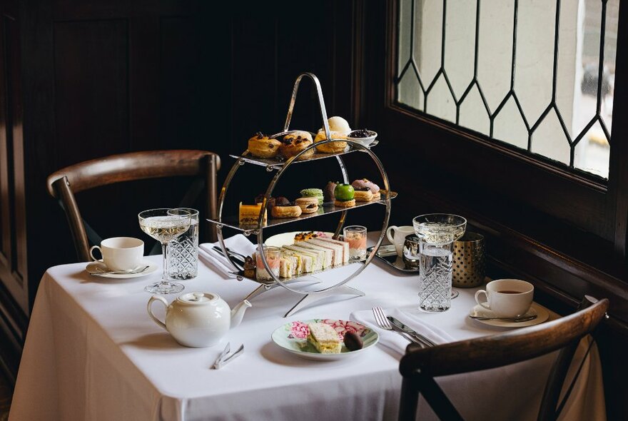 Table with white table cloth and wooden chairs. High tea stand on top is filled with cakes and sandwiches.