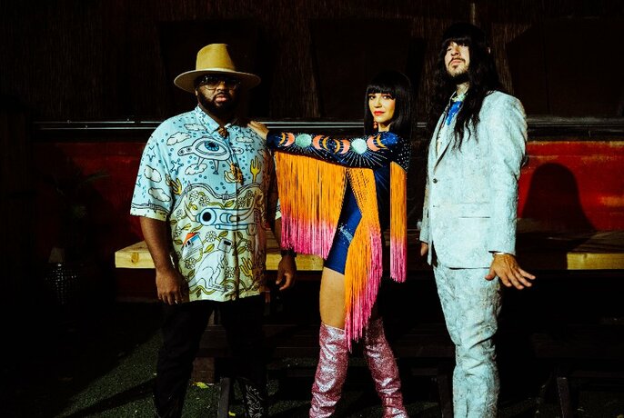 The three members of the band Khruangbin posing in a dark room, the person in the middle wearing a fringed jacket and knee-high boots.