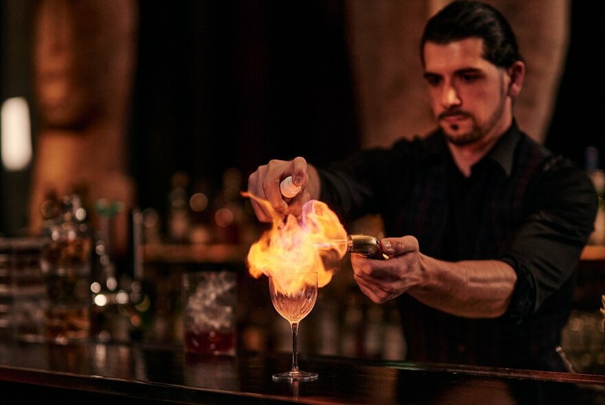 Bartender behind a bar, igniting a cocktail with a firelighter.