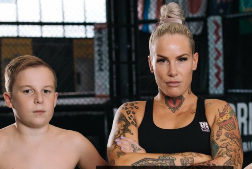Champion bare-knuckle boxer, Bec Rawlings, standing with arms crossed looking directly at the camera, with her shirtless young son standing next to her.