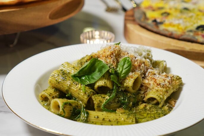 A bowl of rigatoni pasta with a green pesto sauce and basil leaves for garnish.