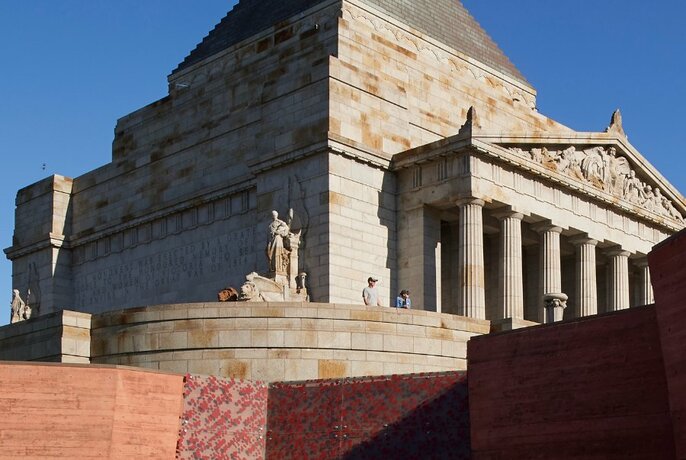 The Shrine of Remembrance, a large concrete monument with a column facade, statues and steps, set against a blue sky.
