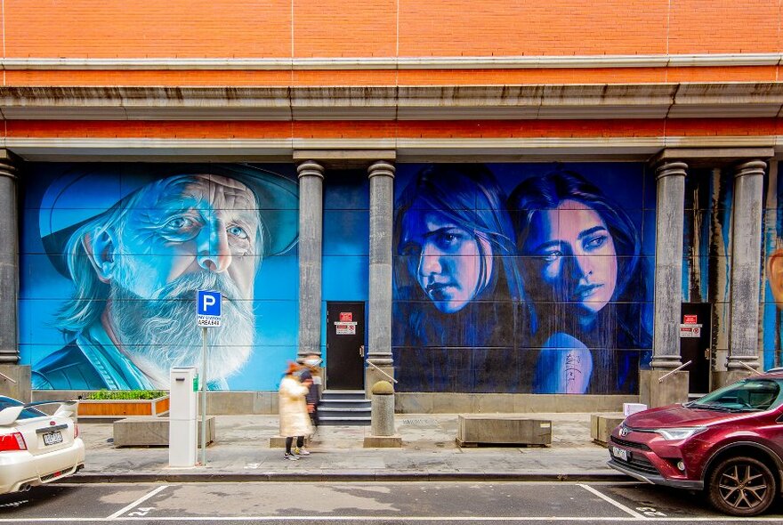 People walking past two blue large-scale street murals, one of a man's face, the other of two young women.