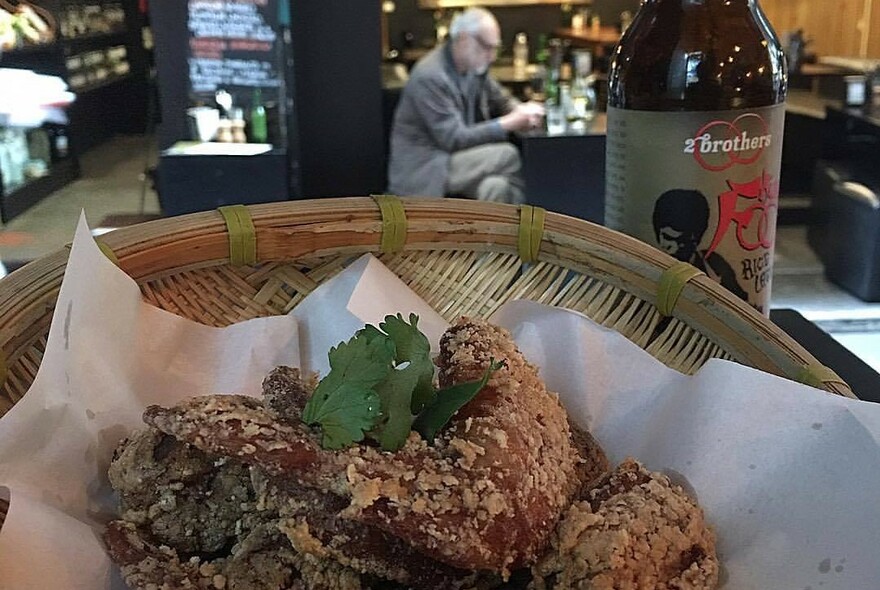Basket with fried dish in foreground, person sitting at a bar with bottle in the background.