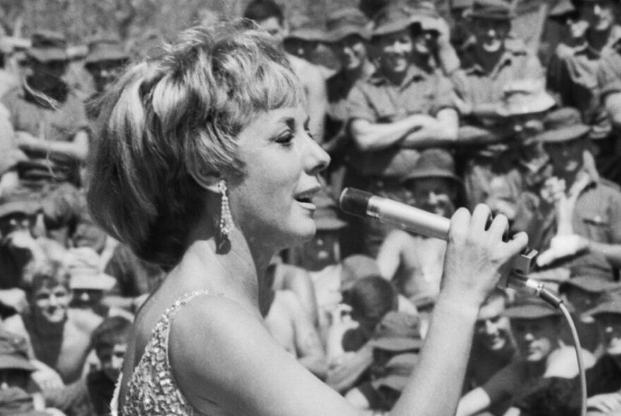 Black and white of woman, seen from side, with bobbed hair singing into thin mic, with soldiers behind.