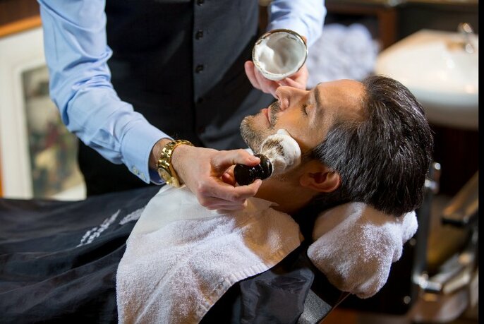 A barber applying shaving cream to the face of a man who is lying back in a barber's chair with a white towel under his chin. 