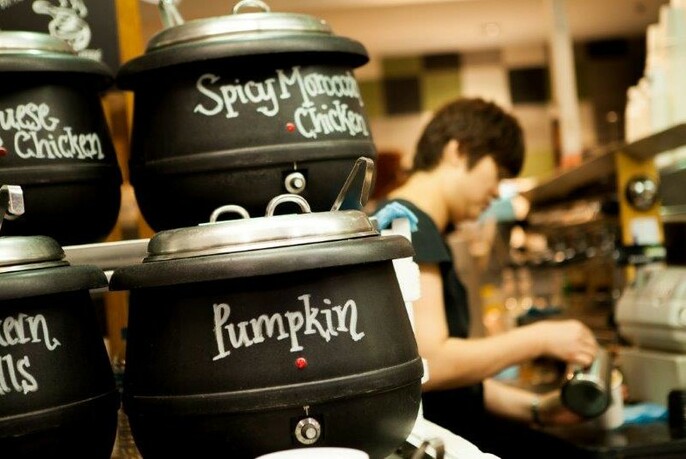 Black pots of pumpkin and chicken soup with lids, with barista pouring coffee.