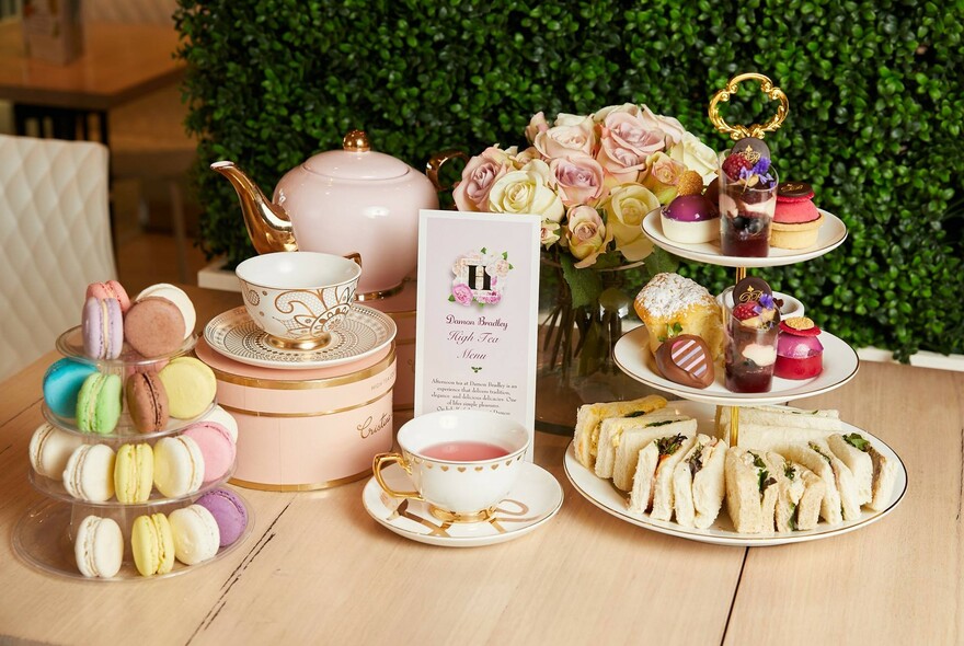 Afternoon tea presentation with cups and saucers, roses, a pink teapot, tier of macarons and cake stand with sandwiches and petits-fours.
