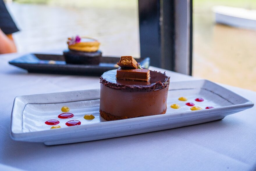 A gourmet chocolate dessert on a white plate with pink and yellow dots of sauce, on a white covered table in front of a window with the water of the Yarra River in the background.