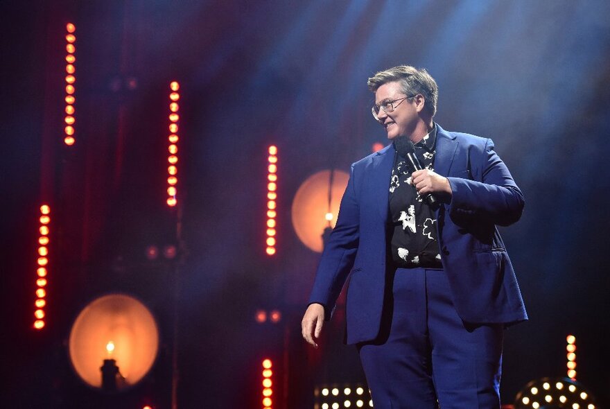 A smiling comedian in a blue suit and glasses, holding a microphone on stage in front of a series of orange lights.