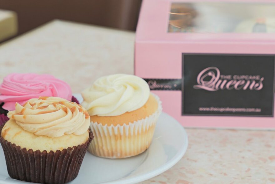 A plate with three icing-topped cupcakes next to a box of branded cupcakes.