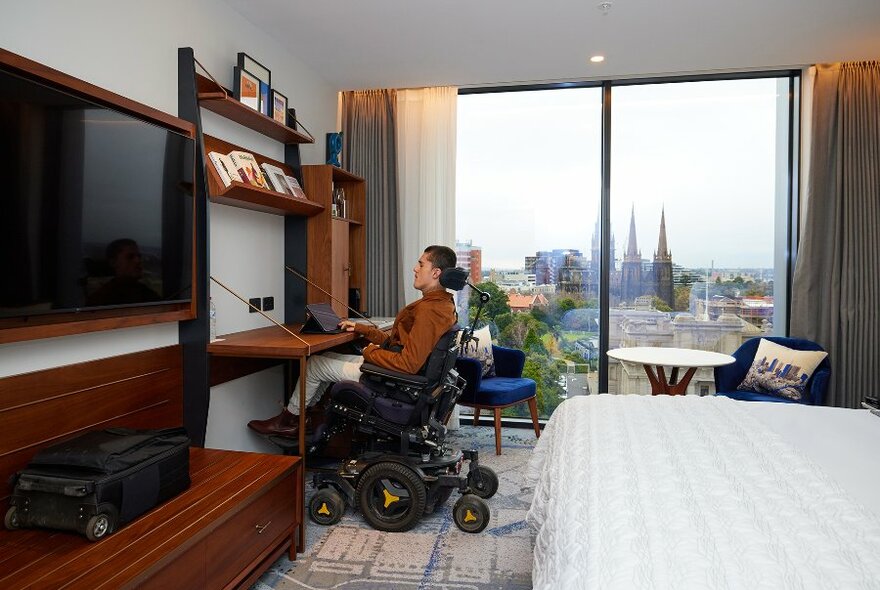 A man sits in a wheelchair at a desk in a hotel room with city views. 