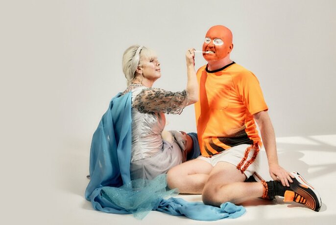 Two people sitting on a floor, both in theatrical costumes, one person applying orange paint on the face of of the other person in front of them.