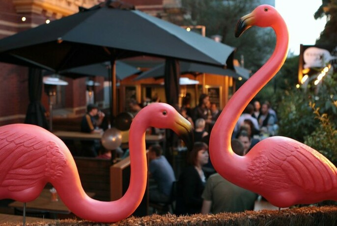 Two pink flamingo statues in front of a busy outdoor bar with black umbrellas.
