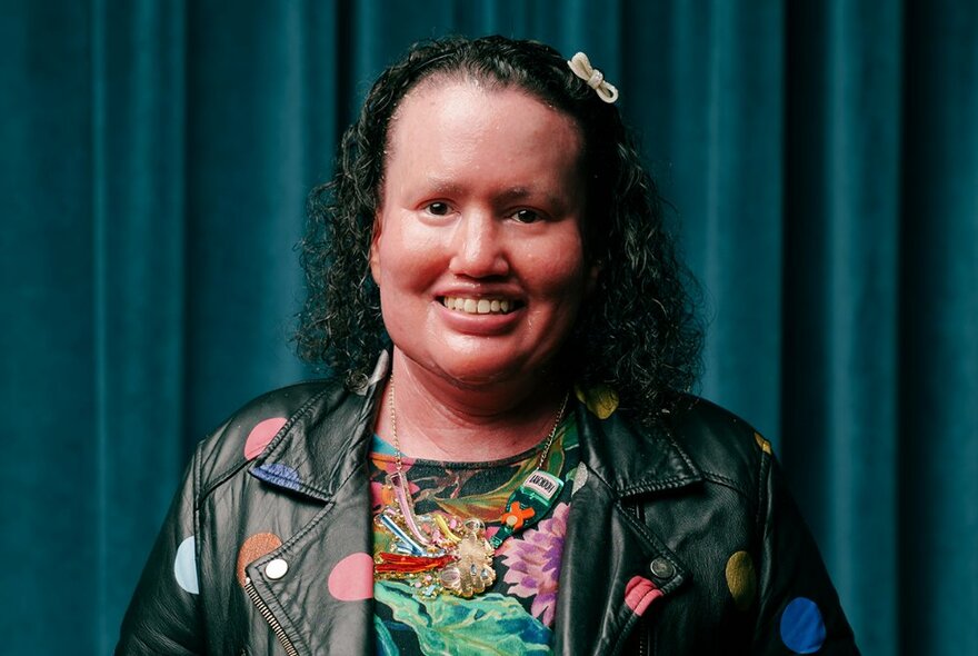 Writer Carly Finlay smiles warmly in front of a green curtain. 