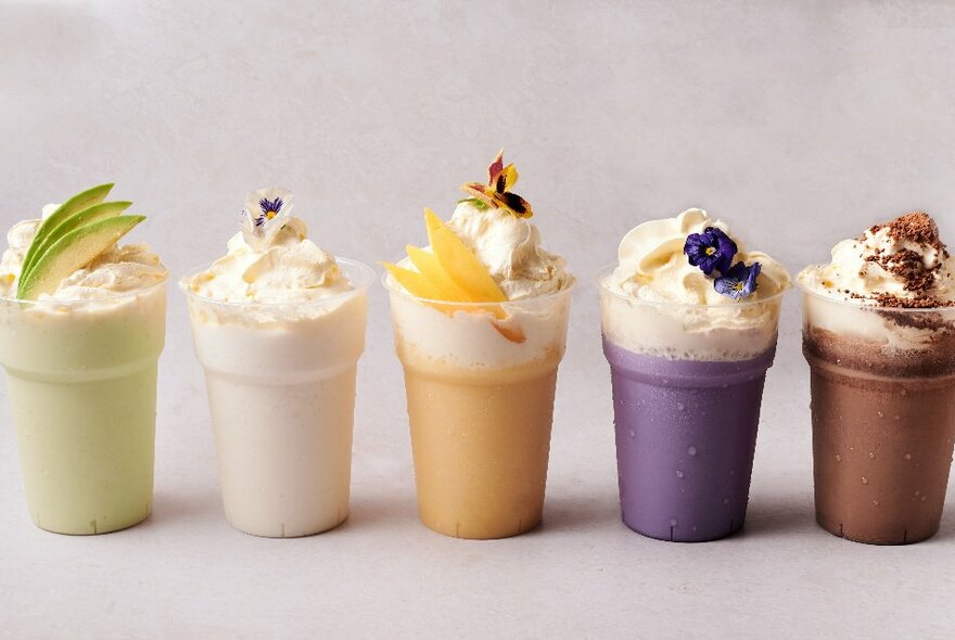 Five different coloured iced drinks or desserts lined-up on a flat surface, all with a creamy topping and fruit.