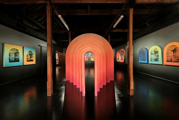 Brightly coloured paintings hanging on dark gallery walls, with a central sculpture of orange cascading arches.