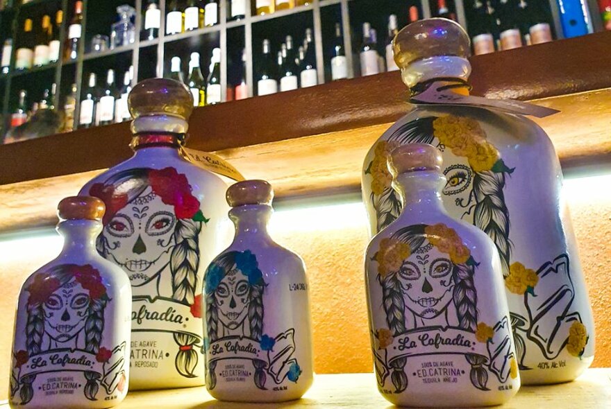 Painted tequila bottles lined up on a bar beneath shelves of wine.
