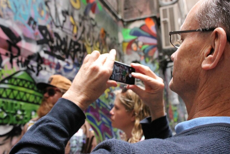 Spectacled man photographing laneway street art with his mobile phone.