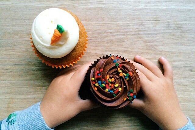 A child's hands holding a chocolate cupcake dotted with coloured sweets, next to a cupcake topped with white icing and a marzipan carrot.
