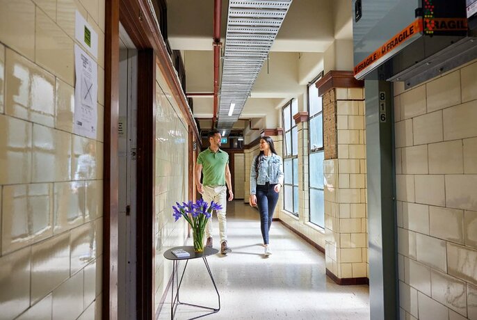 Two people walking down a corridor in the Nicholas Building with tiled walls and sunlight filtering through windows.
