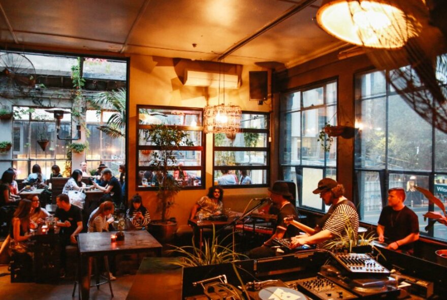 Interior of bar showing a three piece band playing live in front of a mixing desk, people seated at small tables and on couches, and large windows overlooking the street.