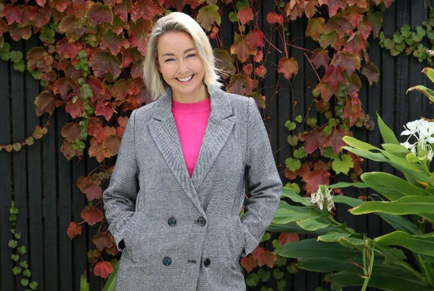 Smiling woman with a blond bob haircut wearing a long grey double-breasted coat, standing in an outdoor garden. 